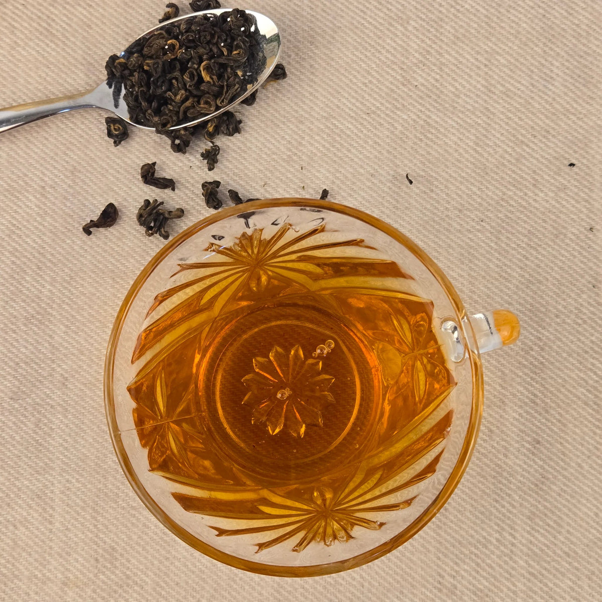 Tea cup with floral design, spoonful of tea leaves, and loose tea leaves on a beige background