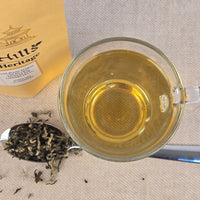Glass of white tea with a spoonful of tea leaves and a box labeled 'Hills Heritage' on a textured surface.